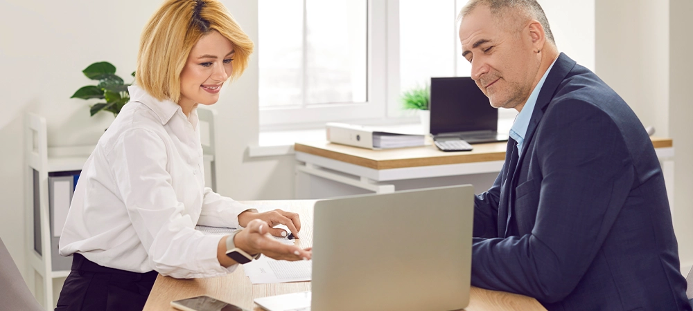 Smiling woman consultant and client sitting at desk using laptop discussing insurance banking services in office. Mature man having consultation with financial advisor, real estate, insurance agent