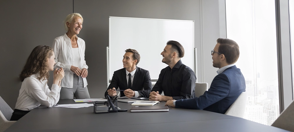 Happy business team having pleasant talk in contemporary board-room. Seminar or corporate training event led by smiling mature 50s business coach, share knowledge, explain strategy to staff members