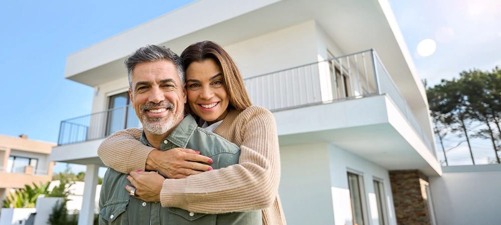 Happy smiling mature older family couple new property buyers modern dream luxury villa owners standing outside house, senior middle aged man and woman in love hugging outdoor.
